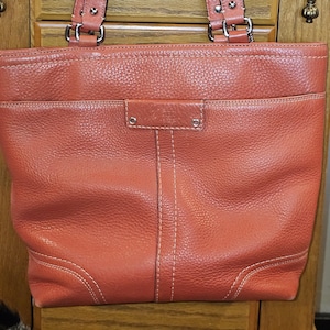May include: A coral-colored leather tote bag with double handles and a front pocket. The bag features white stitching and silver-tone hardware. The Coach logo is visible on the front. The bag is made of textured leather.