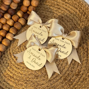 Könnte beinhalten: Drei runde, hölzerne Save-the-Date-Karten mit goldener Beschriftung und einem beigefarbenen Band, das zu einer Schleife gebunden ist. Die Karten lauten "Save the Date Amanda & David" und "Save the Date Amanda David" mit dem Datum "May 06, 2026" auf einer Karte.