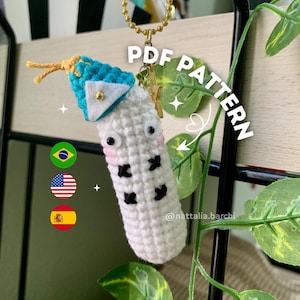 Op de afbeelding: Een handgemaakte witte gehaakte sleutelhanger met zwarte accenten, een blauw-witte bovenkant en een gouden ketting. De sleutelhanger heeft een cartoonachtig ontwerp met een kleine gouden knoop en de tekst "PDF PATTERN". De afbeelding bevat ook de vlaggen van Brazilië, de Verenigde Staten en Spanje.