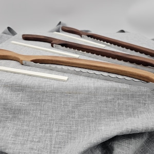 May include: Three wooden bread knives with serrated stainless steel blades are displayed on a grey linen surface. Two knives have dark brown handles, while the other has a lighter wood handle. White rectangular guides are also present.