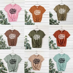 May include: Nine different colored t-shirts, each with the text "Your Design Here" in a script font. The shirts are displayed on hangers against a white wood-paneled background. Colors include pink, brown, olive green, and teal.