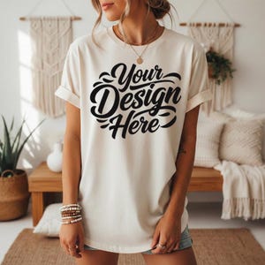 May include: Cream-colored t-shirt with rolled-up sleeves. The shirt features the text "Your Design Here" in a black, stylized font. The shirt is part of a clothing line.