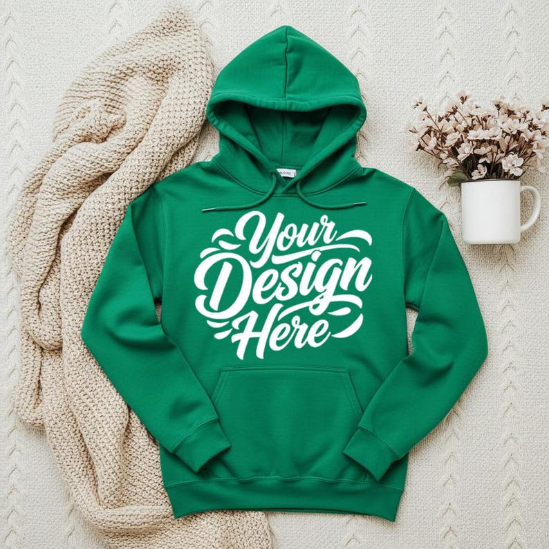 May include: A green hooded sweatshirt with the text "Your Design Here" in white script. The sweatshirt has a front pocket and a drawstring hood. A beige knitted blanket and a white mug with flowers are in the background.