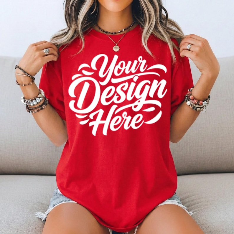 May include: A red t-shirt with the text "Your Design Here" in white script. The shirt is a solid color and appears to be made of a soft material. The model is wearing bracelets and necklaces.