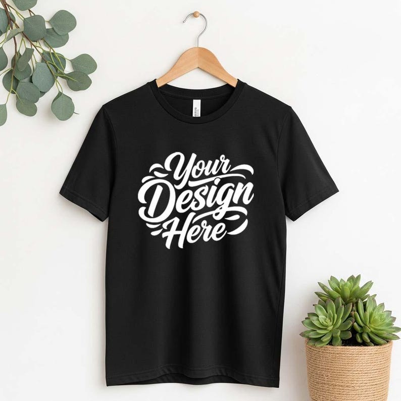 May include: Black crew neck t-shirt with the text "Your Design Here" in white script. The t-shirt is hanging on a wooden hanger against a white background. A small potted plant is visible on the right.