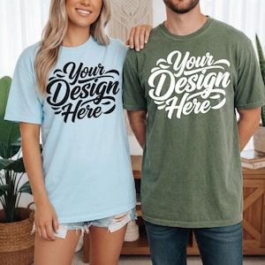 May include: Two short-sleeved t-shirts, one light blue and one olive green, each with the text "Your Design Here" in a stylized script. The shirts are worn by two people, suggesting a matching set or design concept.