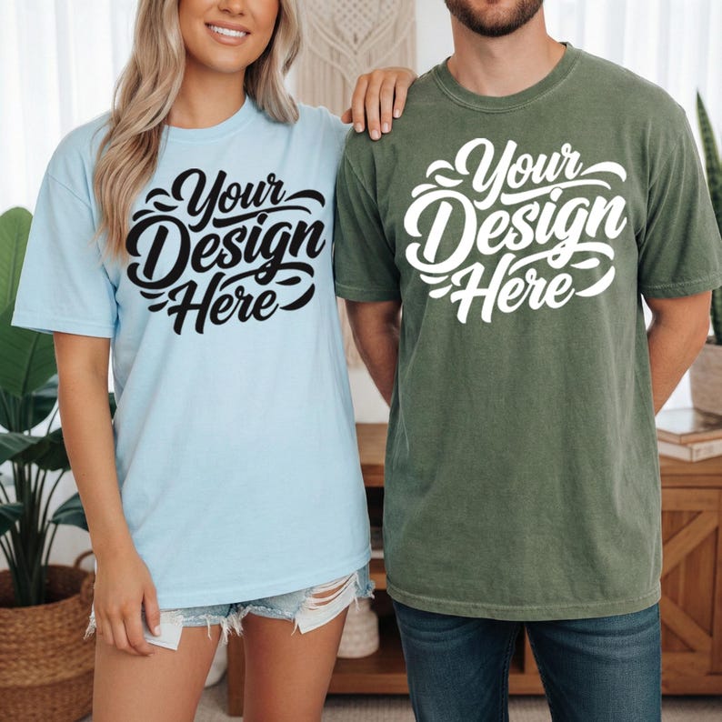 May include: Two short-sleeved t-shirts, one light blue and one olive green, each with the text "Your Design Here" in a stylized script. The shirts are worn by two people, suggesting a matching set or design concept.