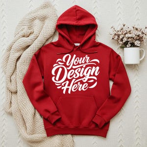 May include: A red hooded sweatshirt with the text "Your Design Here" in white script across the front. The sweatshirt has a front pocket and a drawstring hood. A beige knitted blanket and a white mug with flowers are in the background.