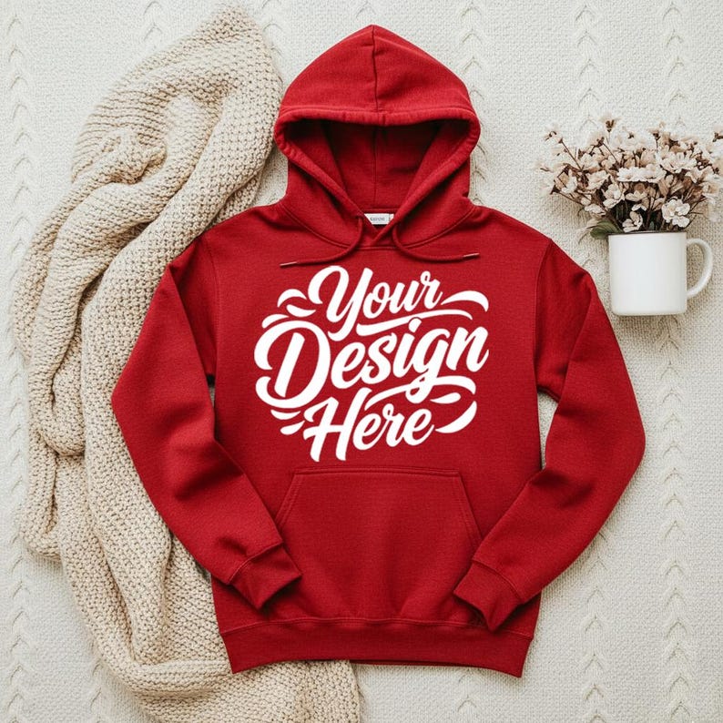 May include: A red hooded sweatshirt with the text "Your Design Here" in white script across the front. The sweatshirt has a front pocket and a drawstring hood. A beige knitted blanket and a white mug with flowers are in the background.