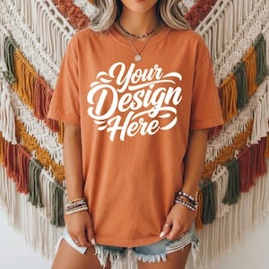 May include: An orange t-shirt with the text "Your Design Here" in white script. The shirt is short-sleeved and appears to be made of a soft material. The model is wearing denim shorts and bracelets.