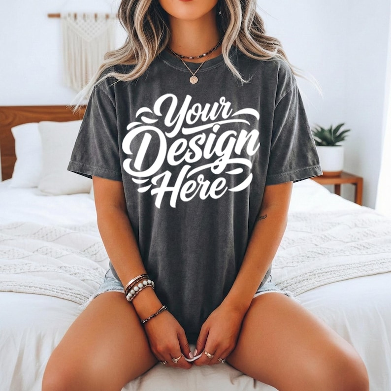 May include: Dark gray t-shirt with the text "Your Design Here" in white script. The shirt is a relaxed fit and is being worn with denim shorts. The model is sitting on a bed with white bedding.