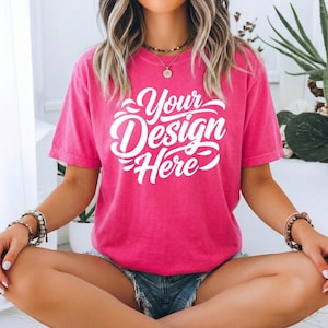 May include: A hot pink t-shirt with the text "Your Design Here" in white script. The short-sleeved shirt is made of a soft material and is being worn by a person. The person is wearing denim shorts and bracelets.