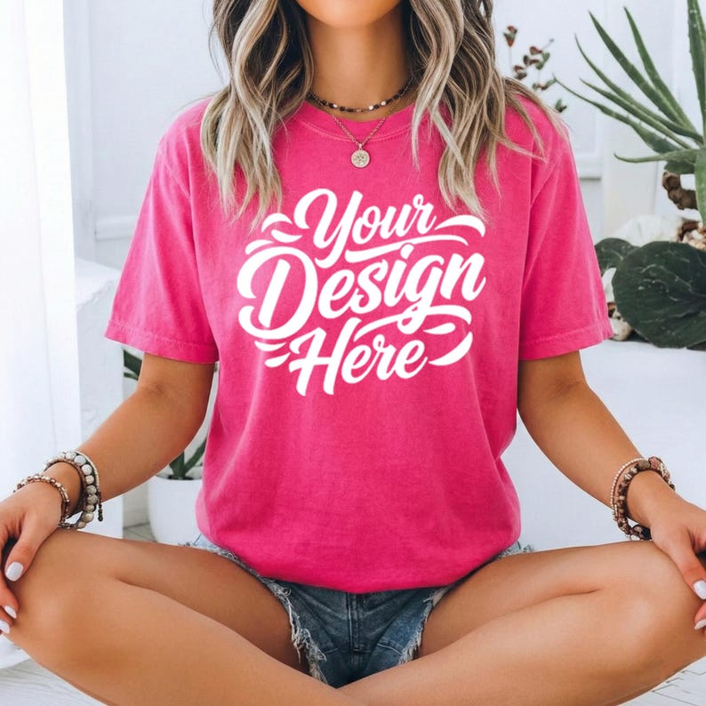 May include: A hot pink t-shirt with the text "Your Design Here" in white script. The short-sleeved shirt is made of a soft material and is being worn by a person. The person is wearing denim shorts and bracelets.