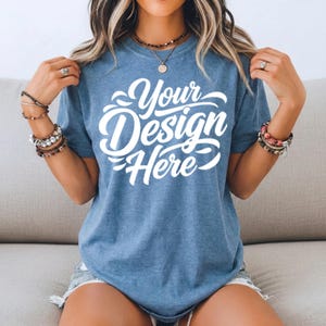 May include: A blue t-shirt with the text "Your Design Here" in white script. The shirt is short-sleeved and has a crew neck. The model is wearing bracelets and rings.