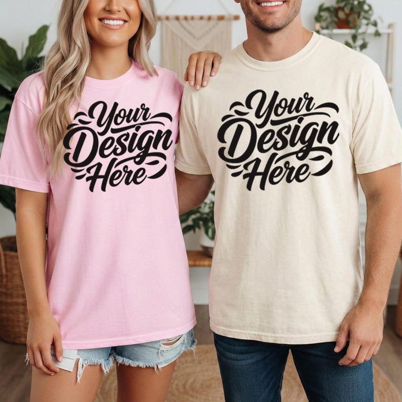 May include: Two short-sleeved t-shirts, one light pink and one cream, each with the text "Your Design Here" in a black, stylized script. The shirts are worn by two people, suggesting a matching set or couple's apparel.