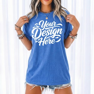 May include: A blue t-shirt with the text "Your Design Here" in white script. The shirt is a solid blue color and has a relaxed fit. The model is wearing denim shorts and jewelry.