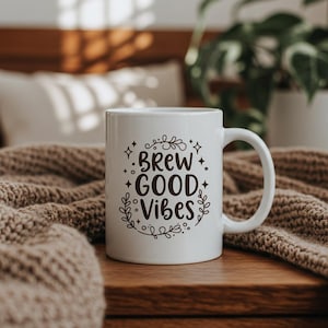 May include: White ceramic coffee mug with black text that reads "Brew Good Vibes" surrounded by a floral design and stars.