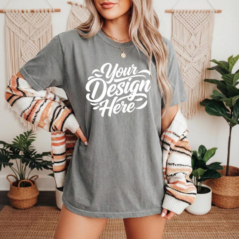 May include: A gray t-shirt with the text "Your Design Here" in white script. The person is wearing a striped sweater over the shoulders. The background includes decorative wall hangings and potted plants.