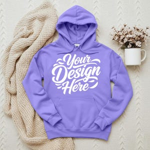 May include: A lavender hoodie with a front pocket and a drawstring hood. The words "Your Design Here" are printed in white script across the front. A beige blanket and a white mug with flowers are in the background.