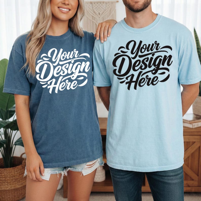 May include: Two short-sleeved t-shirts, one in a faded denim blue and the other in a light blue. Each shirt features the text "Your Design Here" in a stylized script. The shirts are worn by two people.