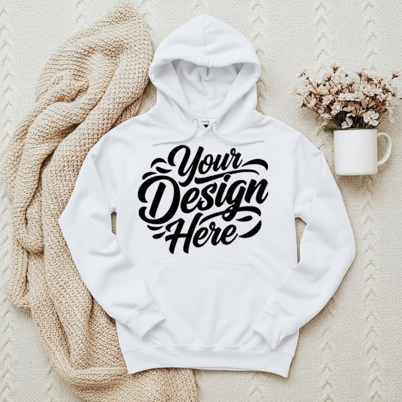 May include: A white hooded sweatshirt with the text "Your Design Here" in a black script. The sweatshirt has a front pocket and a drawstring hood. A beige knitted blanket and a white mug with flowers are in the background.