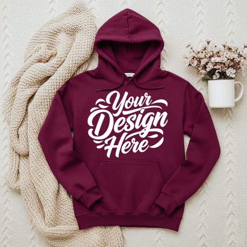 May include: A maroon hoodie with a hood and a front pocket. The hoodie features the white text "Your Design Here" across the chest. A beige knitted blanket is draped on the left side of the hoodie. A white mug with flowers is on the right.