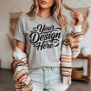 May include: A heather gray t-shirt with the text "Your Design Here" in a black script. The shirt is paired with light blue denim shorts and a striped sweater in shades of orange, cream, and black.