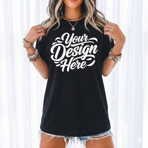May include: Black t-shirt with the text "Your Design Here" in white script. The shirt is a crew neck style and appears to be made of a soft material. The model is wearing denim shorts and jewelry.
