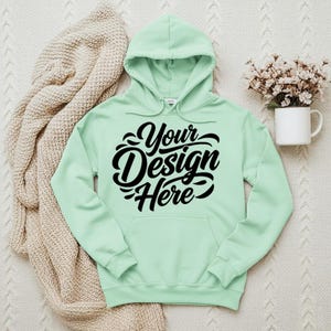 May include: A mint green hoodie with the text "Your Design Here" in black script. The hoodie has a hood and a front pocket. A beige knitted blanket is draped to the left of the hoodie. A white mug with flowers is to the right.