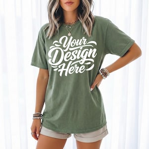 May include: Olive green t-shirt with the text "Your Design Here" in white script. The short-sleeved shirt is worn with tan shorts. The model is wearing bracelets and necklaces.