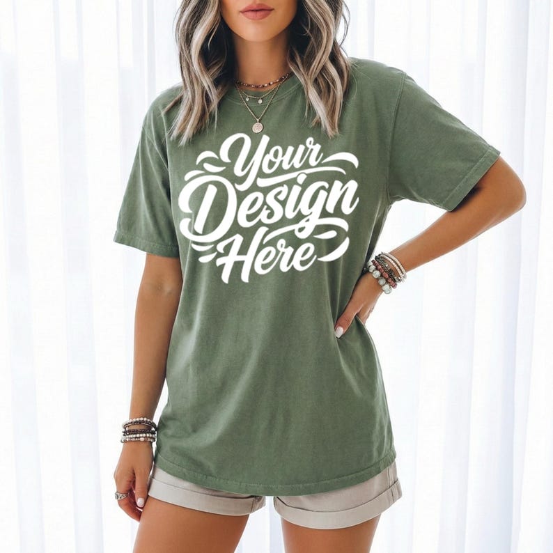 May include: Olive green t-shirt with the text "Your Design Here" in white script. The short-sleeved shirt is worn with tan shorts. The model is wearing bracelets and necklaces.