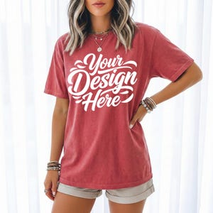 May include: A red t-shirt with the text "Your Design Here" in white script. The shirt has short sleeves and a slightly faded appearance. The model is wearing bracelets and shorts.