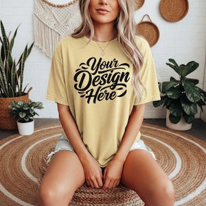 May include: A light yellow t-shirt with the text "Your Design Here" in black script. The shirt is a relaxed fit and is being worn by a person. The person is sitting on a round woven mat.
