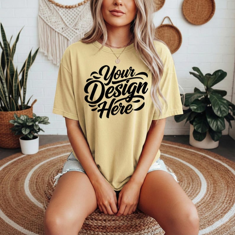 May include: A light yellow t-shirt with the text "Your Design Here" in black script. The shirt is a relaxed fit and is being worn by a person. The person is sitting on a round woven mat.