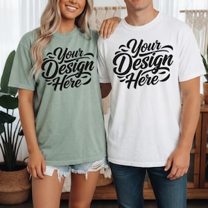 May include: Two people wearing matching t-shirts. One shirt is sage green, the other is white. Both shirts have the text "Your Design Here" in a black, cursive font. The shirts are short-sleeved and appear to be made of a soft material.