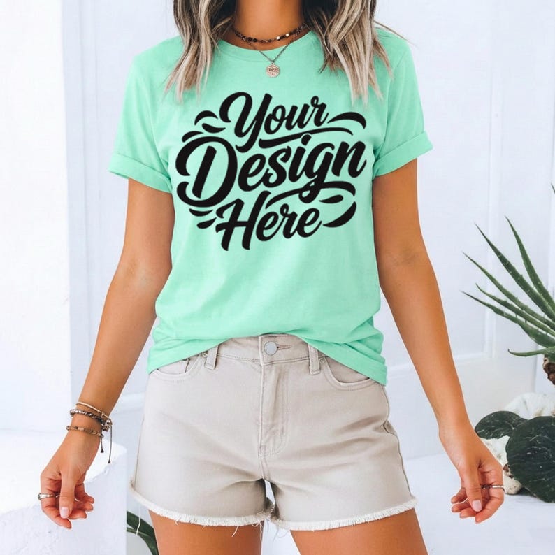 May include: A mint green t-shirt with the text "Your Design Here" in black script. The shirt has rolled-up sleeves and is paired with beige shorts. The overall style is casual and summery.