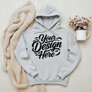 May include: A light gray hoodie with a hood and a front pocket. The hoodie features the text "Your Design Here" in a black, cursive font. A beige knitted blanket and a white mug with flowers are in the background.