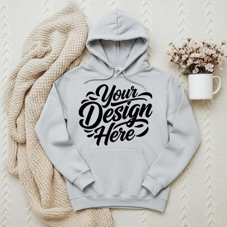 May include: A light gray hoodie with a hood and a front pocket. The hoodie features the text "Your Design Here" in a black, cursive font. A beige knitted blanket and a white mug with flowers are in the background.