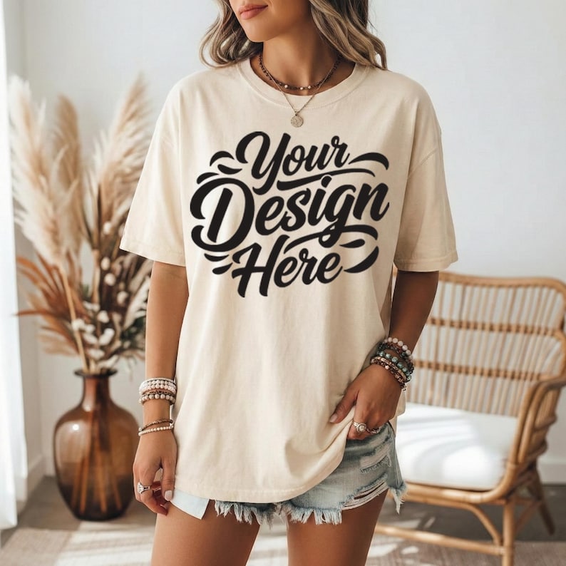 May include: Cream-colored t-shirt with the text "Your Design Here" in black script. The shirt is a relaxed fit and is paired with denim shorts. The model is wearing bracelets and rings.