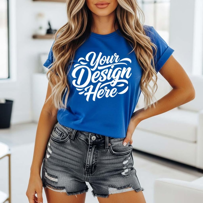 May include: A royal blue t-shirt with rolled sleeves and the text "Your Design Here" in white script. The model is wearing gray denim shorts. The background is a blurred interior.