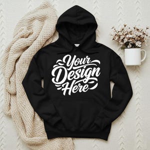 May include: Black hooded sweatshirt with the text "Your Design Here" in white script. The sweatshirt has a front pocket and a hood. A beige blanket and a white mug with flowers are in the background.