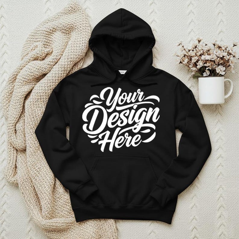 May include: Black hooded sweatshirt with the text "Your Design Here" in white script. The sweatshirt has a front pocket and a hood. A beige blanket and a white mug with flowers are in the background.