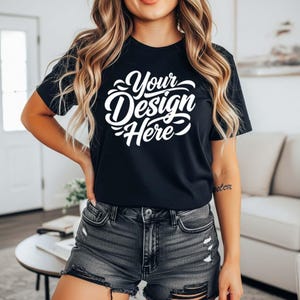 May include: Black crew neck t-shirt with the text "Your Design Here" in white script. The shirt is short-sleeved and appears to be made of a soft material. The model is wearing black denim shorts.