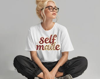 May include: A white t-shirt with the words "self made" in a red and tan leopard print design. The person in the image is wearing black pants and glasses.