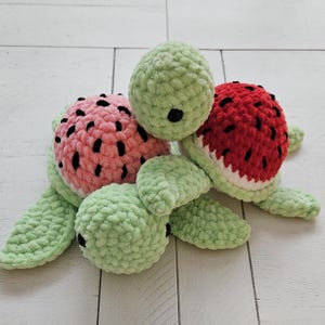 May include: Three plush turtle toys. Two have watermelon-themed shells, one pink with black seeds, the other red with black seeds. The turtles have green bodies and heads, with black eyes. The toys are on a white wooden surface.