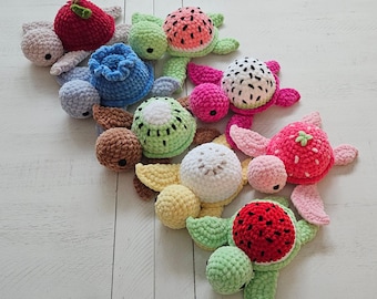 Op de afbeelding: Een verzameling kleurrijke gehaakte fruitturtle. De schildpadden hebben verschillende fruitontwerpen, waaronder een rode appel, een blauwe bosbes, een groene kiwi, een roze drakenvrucht, een rode aardbei en een rode watermeloen. De schildpadden zijn gerangschikt op een wit houten oppervlak.
