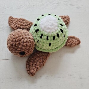 May include: A handmade crochet turtle plushie. The turtle's head is brown, and the shell is green with black spots, resembling a kiwi fruit. The flippers are brown. The plushie is on a white surface.