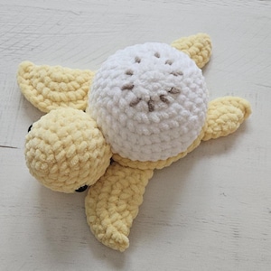 May include: A handmade crochet turtle toy in soft yellow and white yarn. The turtle has a white head with brown embroidered details and yellow flippers and body. The toy is designed with a textured, plush appearance.