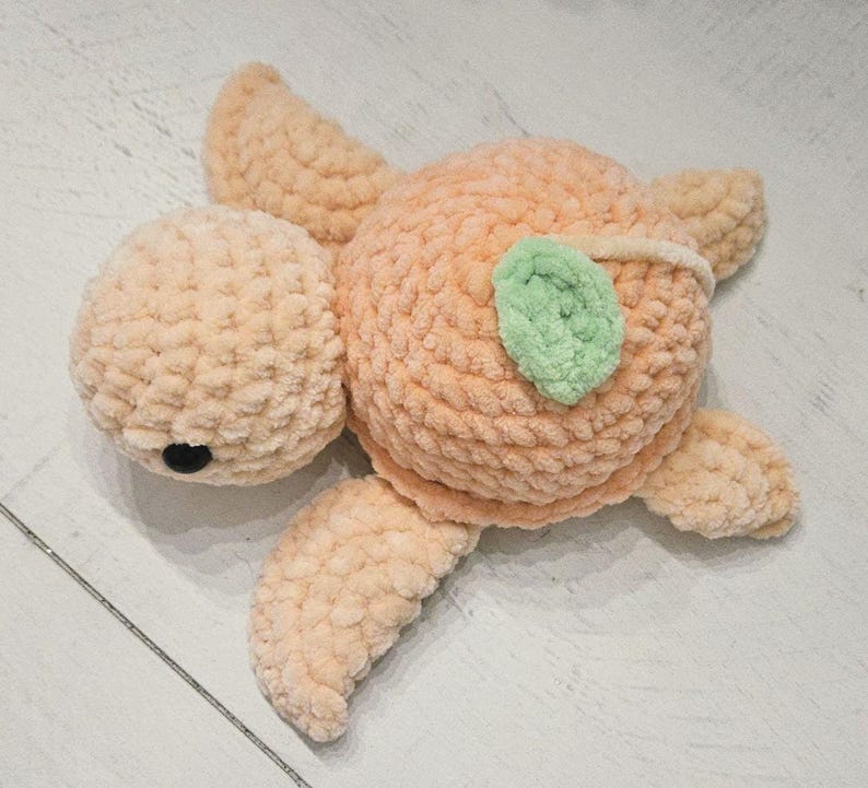 May include: A handmade peach-colored crochet turtle plushie with a green leaf accent. The turtle has a round head, a body, four flipper-like limbs, and a black eye. The plushie is made of soft, textured yarn.