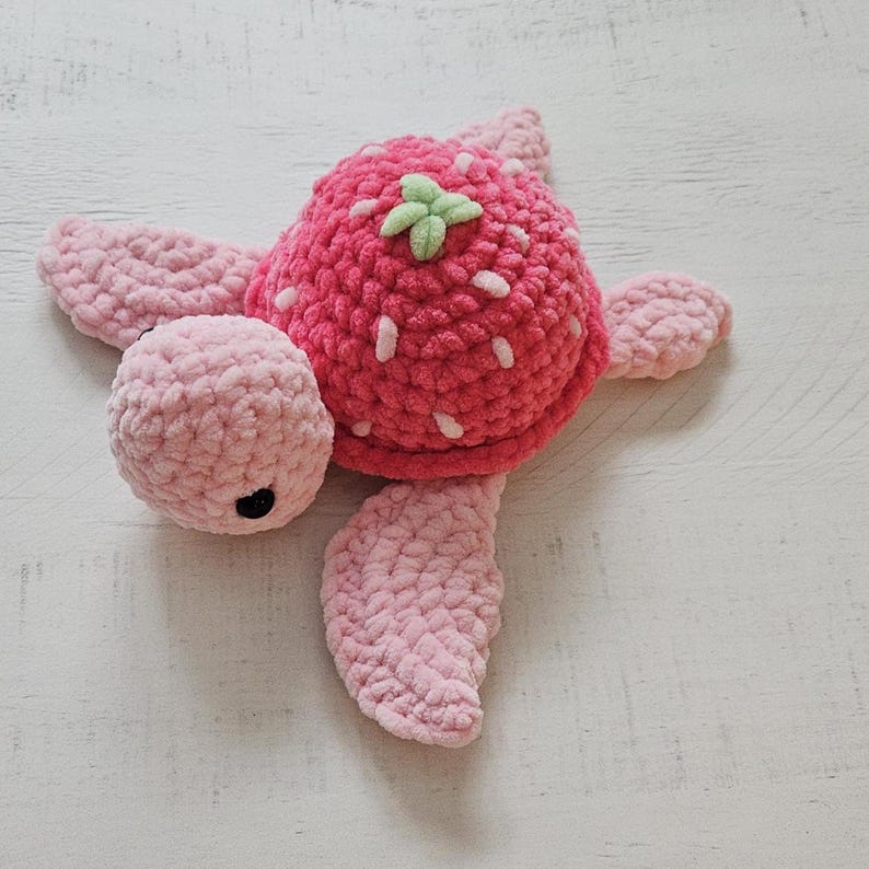 May include: A handmade crochet turtle toy in a strawberry design. The turtle's shell is a vibrant red with white seed details and a green stem. The head, flippers, and tail are a soft pink color. The turtle has a black eye.